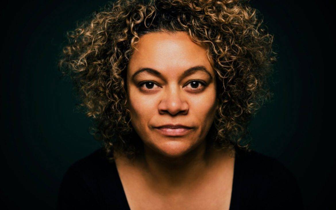 Een lichtgetinte vrouw met bruin halflang krullend haar en een zwart truitje met v-hals