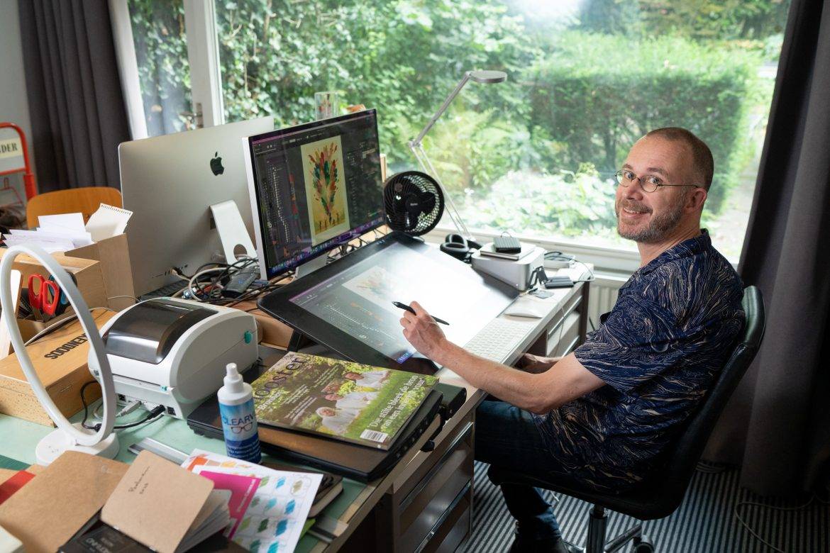 Een man achter een tekencomputer met links op zijn bureau diverse bladen en tekenmateriaal