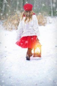 Een meisje met een rode muts, een wit jasje en een rode rok, loopt in de sneeuw met een lamp in haar hand