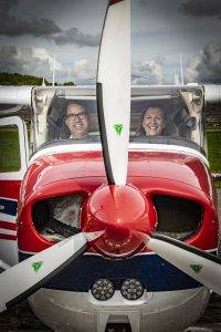 Twee piloten, een man en een vrouw, in de cockpit van een klein vliegtuig. Midden in het beeld de propellor van het vliegtuig.