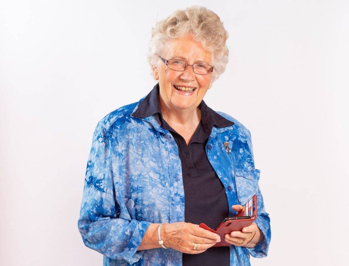 Een oudere dame met grijs haar met krul erin, een blauwe bloese en een witte broek. In haar hand haar telefoon