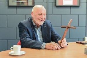 Een man zit aan een houten tafel met een houten kruis in zijn hand. Naast hem staat een witte kop en schotel. Achter hem een betegelde muur.