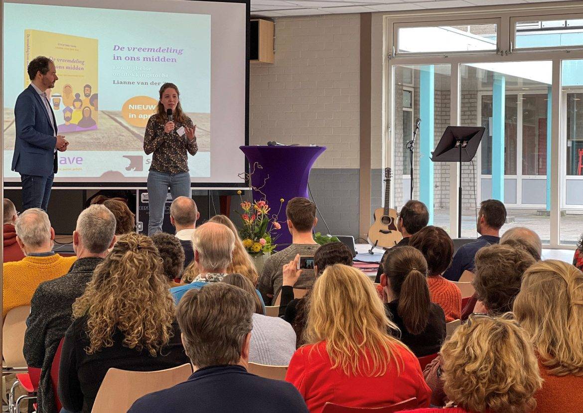 Een jonge vrouw staat met een microfoon in haar hand voor een zaal met mensen. Links van haar staan een man, achter haar hangt een groot scherm met daarop geschreven: De vreemdeling in ons midden