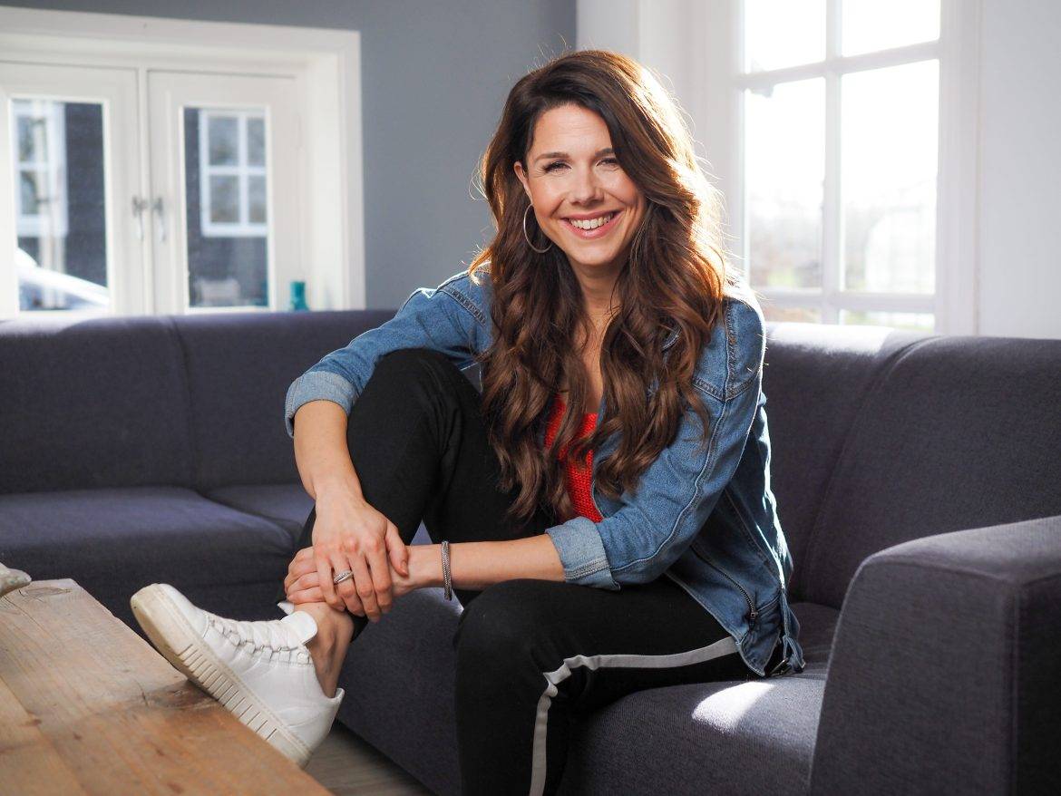 Een vrouw met lang bruin haar en een vriendelijk lachend gezicht zit op een grijze hoekbank met haar witte gymp op de tafel. ze draagt een spijkerblouse en een zwarte joggingbroek met witte streep opzij