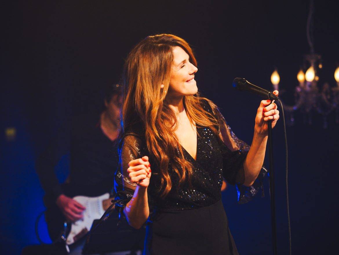 Een zangeres met goudbruin lang haar en een zwarte jurk met v-hals staat voor een microfoon swingend te zingen. Rechts een kroonluchter, links een bassist. Het geheel tegen een donkerblauwe achtergrond