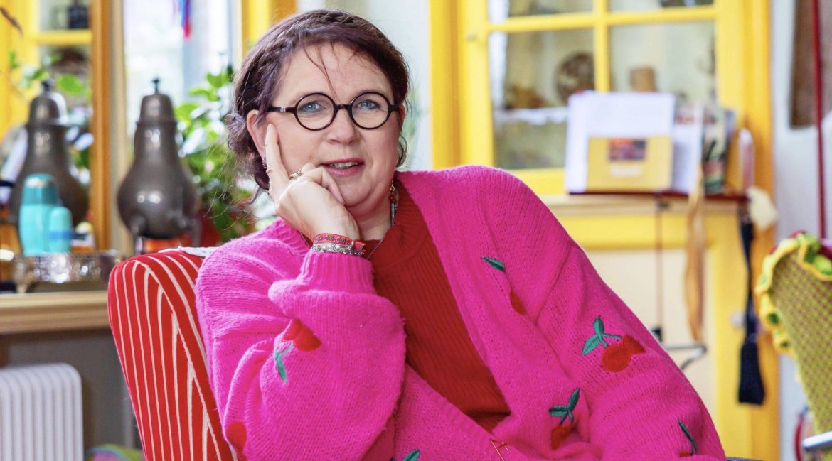 Een vrouw met donker haar en een vlecht over haar hoofd, een bril en een rode trui en broek met roze vest in een rood-wit gestreepte stoel met achter haar een gele kast