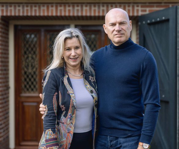 Een stel staat gearmd voor een voordeur. Links de vrouw met blond lang haar en een blauw jasje, rechts de man, met kaal hoofd en blauwe coltrui.