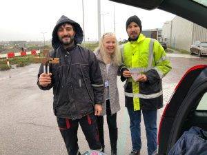 Een vrouw staat buiten tussen twee mannen in werkkleding in. De ene heeft in zijn hand een houten kruis, de ander een boekje.