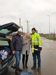 Links een man met een witte helm bij een auto, naast hem een vrouw met lang los haar, rechts van haar een man in gele werkkleding