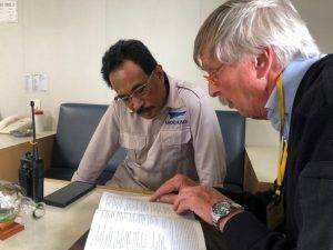 Een man met grijs haar en een keykoord om zijn nek zit naast een man met zwart haar en een bril aan tafel en leest met hem in de Bijbel.