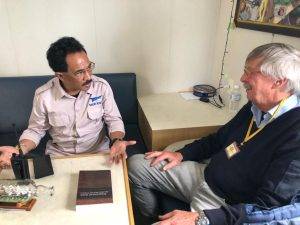 Twee mannen zitten om een tafeltje te praten. Op tafel ligt een boek en een scheepsfles. 