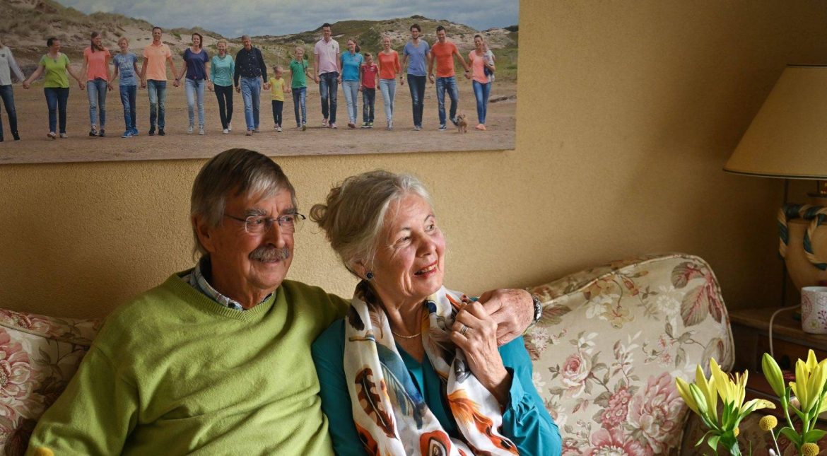 Een echtpaar zit op de bank, hij met de arm over haar schouder. Links op de grond staat een geel boeket. Achter hen hangt een foto aan de wand van een groot gezin op het strand.