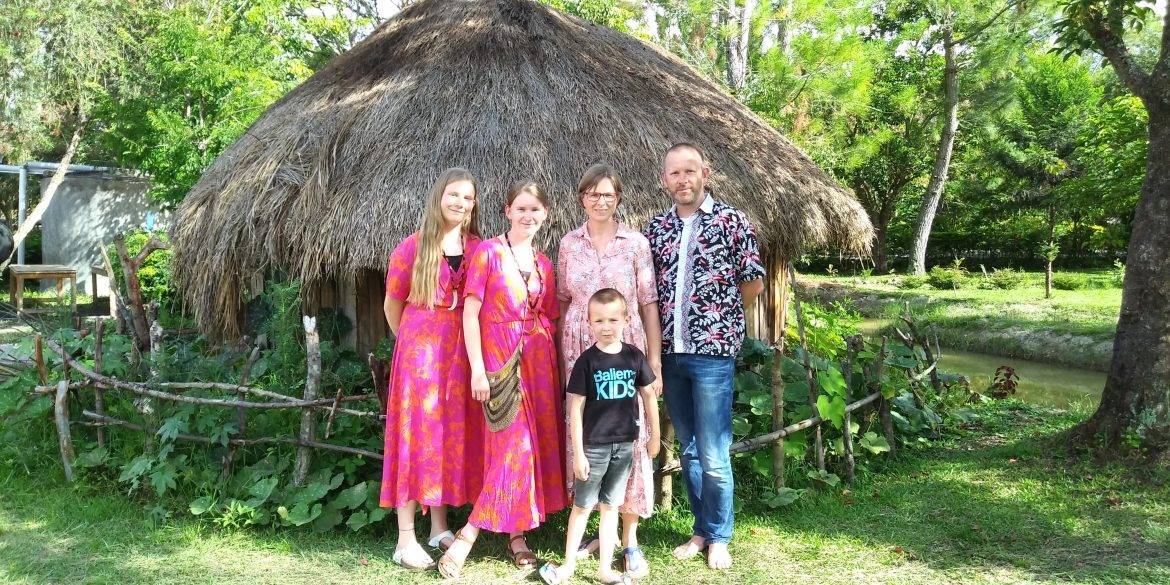 Een man en een vrouw en twee meiden en een jongetje staan voor een ronde hut met een rieten dak. De meisjes dragen felroze lange jurken. Om het gezin heen staan bomen.