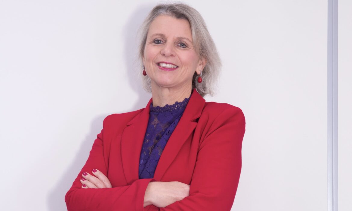 Een vrouw met lichtblond/grijs halflang haar, rode oorbellen, een rood jasje en een blauwe blouse staat met haar armen over elkaar tegen een witte wand