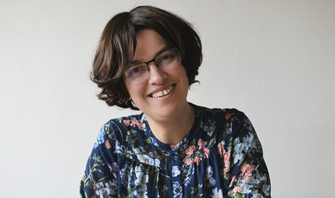 Een vrouw met halflang bruin haar en een bril en een donkerblauwe bloemetjes blouse kijkt lachend en met een scheef hoofd in de camera