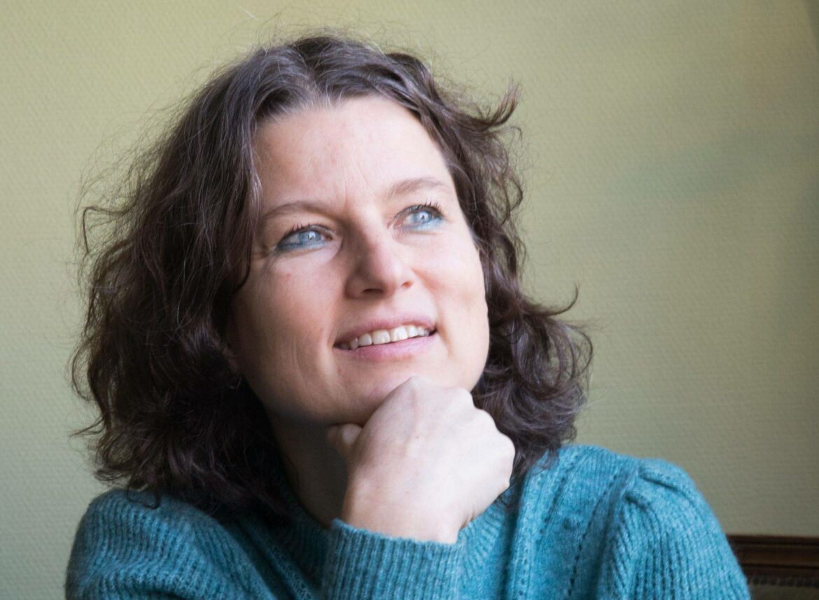 Een vrouw met bruin haar en blauwe ogen en een blauwe trui kijkt lachend omhoog met haar hand onder haar kin