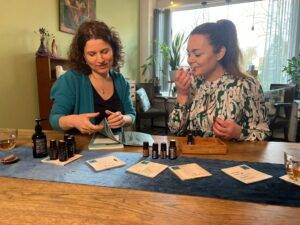 Twee vrouwen zitten aan een tafel. Voor hen staan flesjes olie. De vrouw links bladert in een boekje, de vrouw rechts ruikt aan een glas